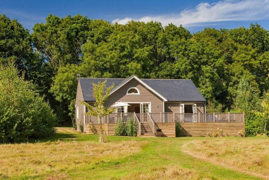 Timber-clad lodge with slate roof and wraparound deck set in meadowland with mature trees behind