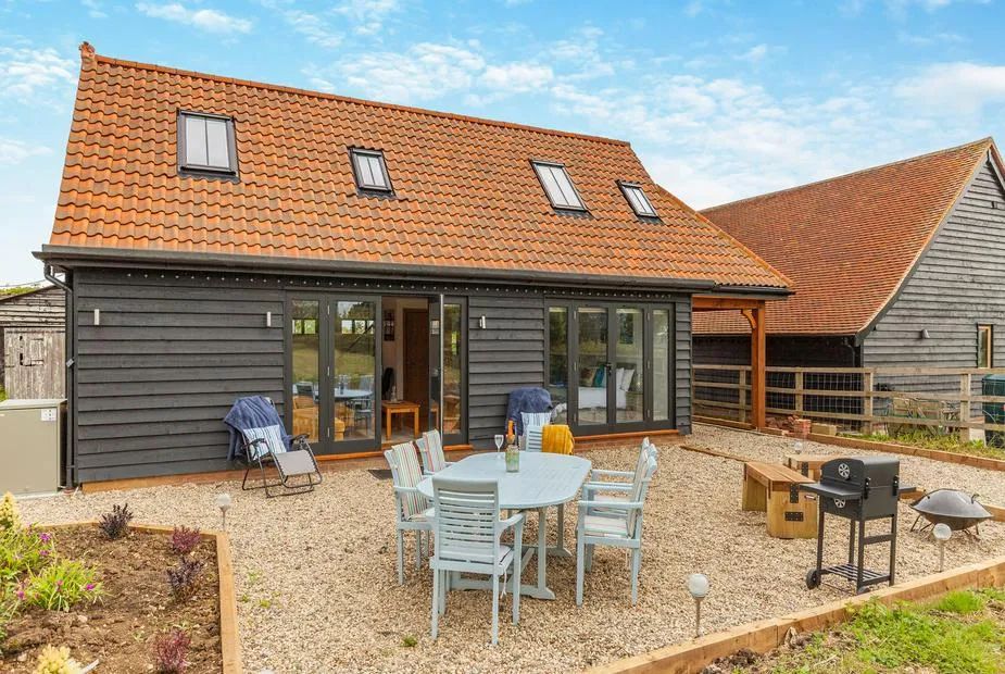 Black timber-clad barn conversion with gravel garden, pale blue dining table, BBQ and sun loungers