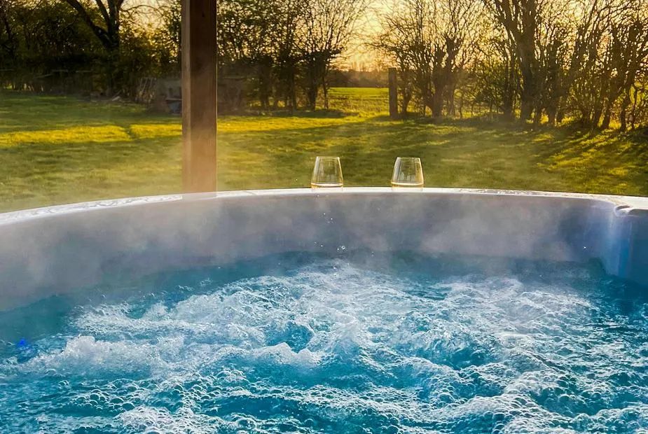 Wood-fired hot tub with two wine glasses on the rim and steam rising over open fields at sunset