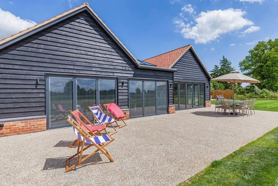 Black timber-clad barn with row of glass doors, gravel patio with striped deckchairs and parasol dining set